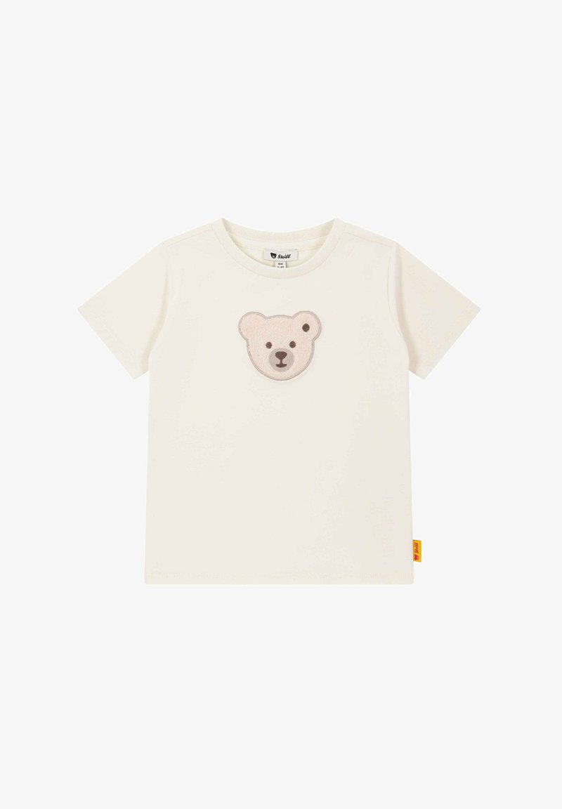 Kremowa bawełniana koszulka z krótkim rękawem, z miękką, haftowaną naszywką w kształcie twarzy misia w odcieniach jasnobrązowym i ciemniejszym brązowym na piersi. Okrągły dekolt.