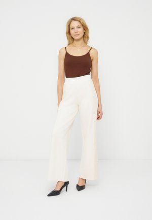 Femme debout portant un pantalon blanc taille haute à jambes larges, un haut marron sans manches, des talons noirs et un collier pendentif, sur un fond blanc uni.