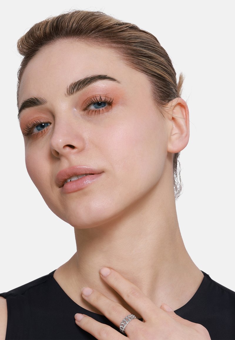 Primo piano di una persona con pelle liscia, trucco neutro e capelli castano chiaro. Indossa un anello a catena in argento, una maglia nera e mostra una posa con la mano.