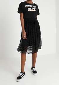 Musta t-paita, jossa on "DESERT DAZE" -printti, yhdistetty läpikuultavaan mustaan pilkulliseen hameeseen. Mustat tennarit viimeistelevät asun.