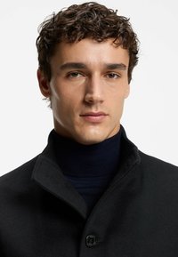 Cappotto di lana nera con colletto alto e chiusura a un bottone, indossato sopra un dolcevita blu scuro. In primo piano, il viso, che mostra un incarnato chiaro e capelli ricci.