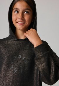Sudadera con capucha negra y brillo metálico, que presenta un logo redondo "SAINT" en el centro. Los puños y el dobladillo acanalados añaden textura.