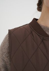 Brun vadderad väst med diamantsmönster och räfflad krage, kombinerad med en ljusbrun texturerad tröja. Fokus på tyg- och snittdetaljer.