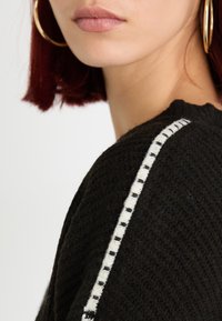 Maglione nero con texture, caratterizzato da una striscia bianca a motivo lungo la spalla. Il tessuto sembra lavorato a maglia con una finitura morbida.