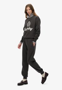 Vrouw in een donkergrijze sweatshirt en joggingsbroek, gecombineerd met een gestreept shirt en kastanjebruine instappers, loopt tegen een witte achtergrond.