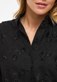 Svart broderad skjorta med blommönster, med krage, knäppning fram och texturerad tyg. Broderiet är tonalt och detaljerat.