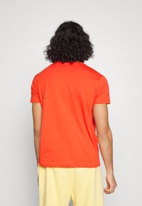 Rotes T-Shirt mit kurzen Ärmeln aus glatter Baumwolle, mit Rundhalsausschnitt und geradem Schnitt, getragen von einer Person mit lockigem Haar.