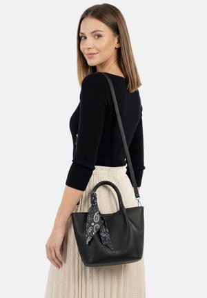 Jonge vrouw met zwarte top en lichte polkadot rok, draagt zwarte leren handtas met zwarte patterned sjaal.