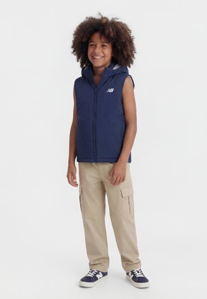 Naeratav laps lokkis juustega, kellel on seljas tumesinine varrukateta kapuutsiga dressipluus, beežid töömugavad püksid ja tumesinised tossud valgete talladega.