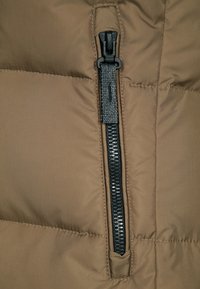 Detailní záběr na černý zip s látkovým taháčkem na hnědé prošívané bundě, ukazující detaily šití.