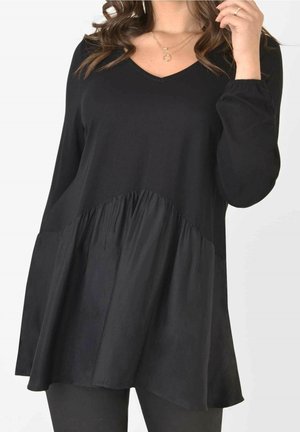 Vrouw draagt een zwarte lange mouw V-hals tuniek met geplooide details in de taille en zwarte leggings, gezien van de nek tot halverwege het dijbeen.