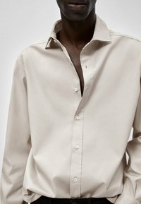 Beżowa koszula z długim rękawem wykonana z gładkiej bawełny, z kołnierzykiem w szpic, widocznymi guzikami oraz rozpiętym klinem dla luźniejszego kroju.