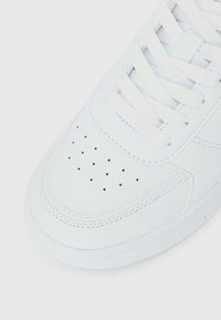 Λευκό δερμάτινο αθλητικό παπούτσι με υφή στην επάνω επιφάνεια, στρογγυλεμένη μύτη, διάτρητες λεπτομέρειες και επίπεδα λευκά κορδόνια. Λείο πέλμα, με ελάχιστες ραφές ως διακοσμητικά στοιχεία.