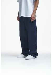 Pantalones de mezclilla azul oscuro con un corte holgado, con costuras visibles y un diseño clásico, combinados con zapatillas blancas sobre un fondo liso.