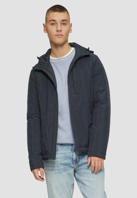 Marineblaue Steppjacke mit Reißverschluss vorne, verstellbare Kapuze und Seitentaschen. Tragend über einem hellblauen Pullover und hellblauen Jeans.