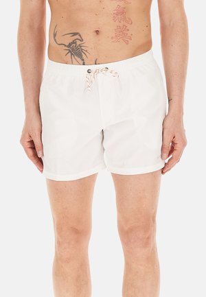 Torace e gambe maschili che indossano pantaloncini da bagno bianchi con coulisse, mostrando tatuaggi sull'addome e sulla parte superiore del petto.