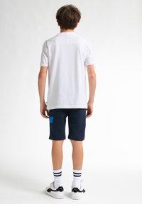 Cămașă albă cu mânecă scurtă, asortată cu șorturi bleumarin cu o pată de accent albastră; completată cu adidași albaștri și negri și șosete cu dungi.