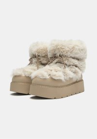 Beige platform csizmák puha szőrmével borítva, felgombolyított szőrme gallérral, fűzős elülső résszel és texturált talppal a tapadás érdekében.