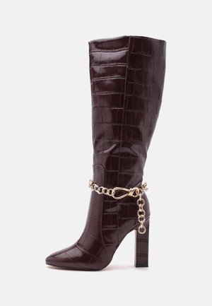Botte brillante brun foncé montant jusqu'au genou avec texture crocodile, bout pointu, talon bloc et détail de chaîne dorée autour de la cheville.