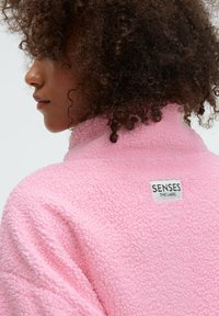 Rosa Fleecejacke mit hohem Kragen, aus weichem, strukturiertem Stoff, mit einem angenähten Etikett mit der Aufschrift "SENSES THE LABEL."