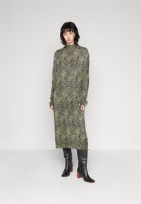 Robe longue ajustée de couleur vert foncé avec un motif léopard, dotée de manches longues et d'un col haut. Portée avec des bottes à talons noires.