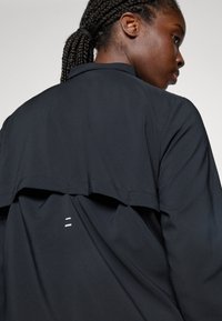 Frau mit geflochtenem Haar, die von hinten und von der Seite in einer schwarzen Jacke gezeigt wird, wobei das belüftete obere Rückenteil und die reflektierenden Details der Jacke hervorgehoben werden.