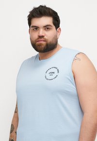 T-shirt sans manches en coton bleu clair avec un design de logo circulaire sur la poitrine. Texture douce, coupe décontractée, col rond simple.