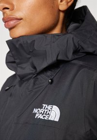Schwarze Windjacke mit hohem Kragen und einem weißen Logo. Hergestellt aus glattem, leichtem Material mit verstellbaren Kordelzügen.
