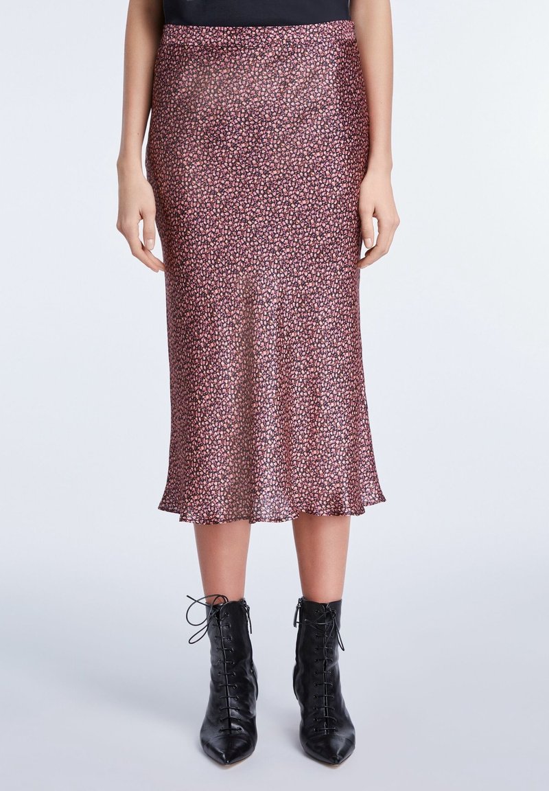 Femme portant une jupe midi fleurie rose et noire, associée à des bottines noires à lacets et bouts pointus, debout devant un fond uni.