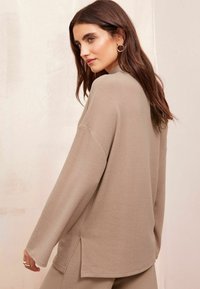 Beige long-sleeve top with a relaxed fit, featuring side slits and a soft texture. Paired with matching bottoms, it has a minimal design.