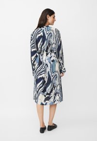 Langes, figurbetontes Kleid mit einem blau-creamfarbenen Marmor-Muster, langen Ärmeln, einem Gürtel in der Taille und einem geraden Saum. Getragen mit schwarzen Ballerinas.