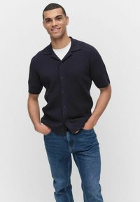 Marineblauw overhemd met korte mouwen, knoopsluiting en een textured patroon, gecombineerd met blauwe jeans. Het overhemd heeft een kraag en zes knopen aan de voorkant.