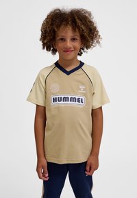 Dziecko z kręconymi włosami w beżowej sportowej koszulce hummel i granatowych spodniach, stojące na tle jasnego, jednolitego tła.