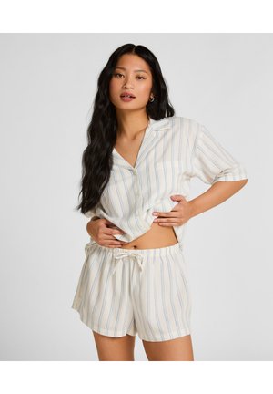 Femme portant une chemise à manches courtes à rayures crème et bleu clair avec des boutons et un short assorti à cordon, posant les mains sur la taille.