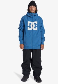 Hoodie zippé bleu avec logo blanc sur la poitrine, associé à un pantalon noir ample et des chaussures noires. Le tissu semble lisse, au design décontracté.