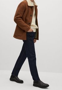 Brun suedet jakke med en blød krave, cremet striktrøje under, mørk denim bukser og sorte Chelsea-støvler. Glat tekstur, afslappet pasform.