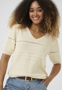 Maglione lavorato a maglia crema con scollatura a V, maniche corte e righe orizzontali testurizzate. Abbinato a jeans in denim e una cintura marrone.