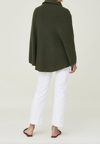 Poncho lavorato a maglia verde oliva con collo alto e maniche larghe, abbinato a pantaloni bianchi a gamba dritta e sandali bassi marroni.