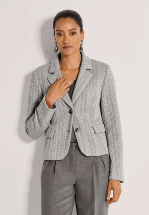Frau trägt einen grauen Kabelstrick-Blazer mit zwei Knöpfen und passenden grauen Hosen, steht vor einem schlichten hellen Hintergrund.