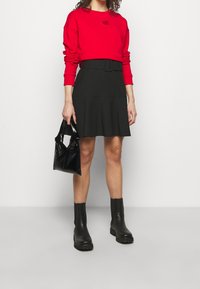 Sudadera roja con parche de logo, falda negra acampanada con cinturón, bolso negro y botas negras de tobillo gruesas. Texturas simples y líneas limpias.