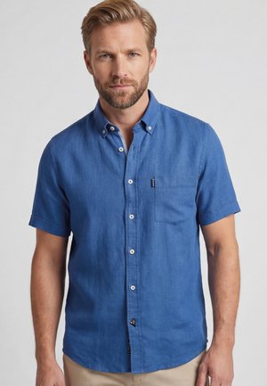 Man met baard die een blauw, kort mouw, knoopjeshemd draagt, staat voor een effen lichtgrijze achtergrond.