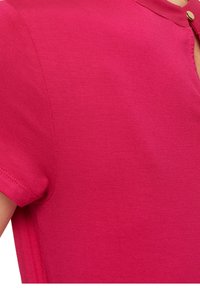 Fuksjowa bluzka z krótkim rękawem, wykonana z miękkiego, gładkiego materiału; posiada okrągły dekolt z małym guzikiem po boku.