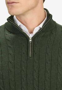 Donkergroene gebreide zip-up trui met kabelpatroon, hoge kraag en zilveren ritsdetail, gedragen over een witte overhemd met kraag.