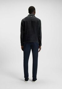 Zwarte suede jas met een gladde textuur, details op de rugnaad en een kraag, gecombineerd met marineblauwe broeken en donkere sneakers.
