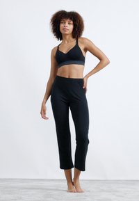 Črn športni nedrček s širokim pasom in usklajenimi črnimi kratkimi leggins hlačami. Tkanina se zdi mehka z gladko teksturo. Model stoji bosa.