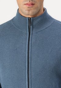 Light blue knitted jacket featuring a high collar, a front zipper, and textured fabric with a ribbed pattern along the neck and body.
