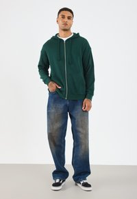 Sweat à capuche zippé vert avec cordon de serrage, deux poches avant, associé à un jean ample bleu clair et des baskets noires avec semelles blanches.