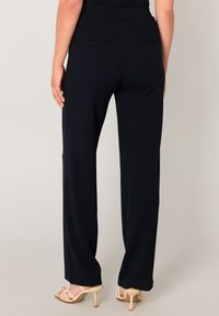 Marineblaue Hosen mit weitem Bein und glatter Textur, ausgestattet mit zwei Gesäßtaschen und dezenten Seitenschlitzen an den Säumen, kombiniert mit beigen Sandalen mit hohem Absatz.