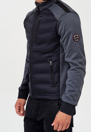 Mężczyzna w czarnej pikowanej kurtce na zamek, z ciemnoszarymi rękawami i okrągłą naszywką na górnej części lewego ramienia, stojący na jednolitym tle.