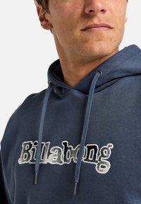 Navy blue hoodie with a drawstring and a textured "Billabong" logo in white and black. The fabric is soft, with a relaxed fit and smooth texture.